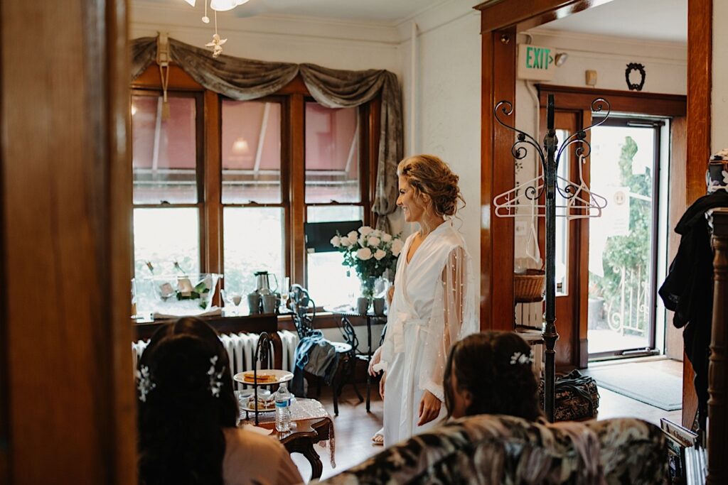 A bride stands in the getting ready space the morning of her wedding day.  She has her hair in a loose updo and she has a white robe on with pearls on the sleeves.