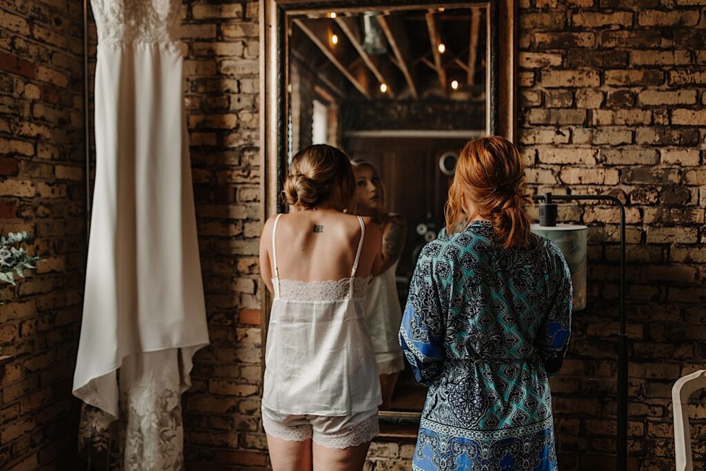 A bride gets ready for her wedding while looking in the mirror as her bridesmaid looks on.