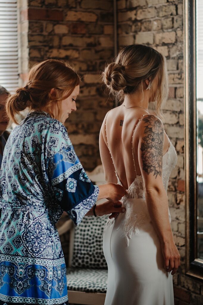 A bridesmaid helps a bride put her wedding dress on while standing in front of a mirror.