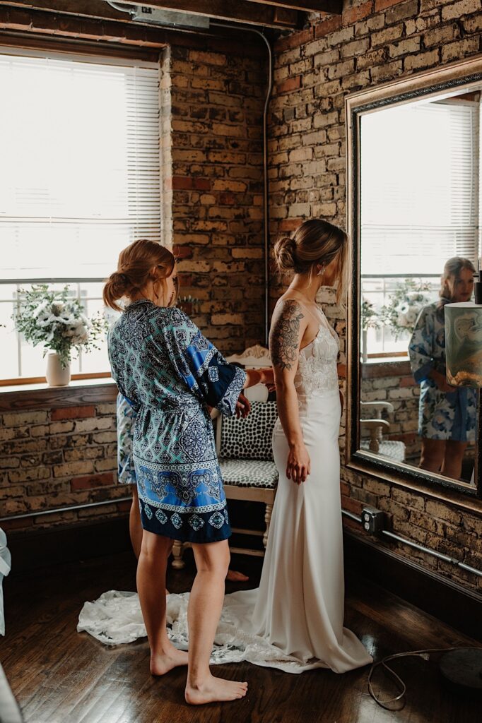 A bridesmaid helps a bride put her wedding dress on while standing in front of a mirror.
