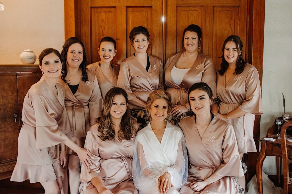 A bride and her bridesmaids all sit together after getting ready for the wedding day in a Chicago Inn.  The bride wears a white robe with pearls and the bridesmaids wear champagne colored robes.
