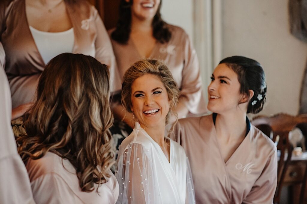 A bride sits in a white robe with pearls on the sleeves next to her bridesmaids that all wear matching pink robes with their initials.
