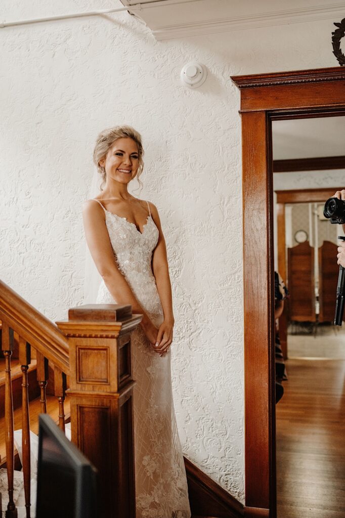 A bride stands at the end of a staircase with her hands folded in front of her smiling directly at the camera.