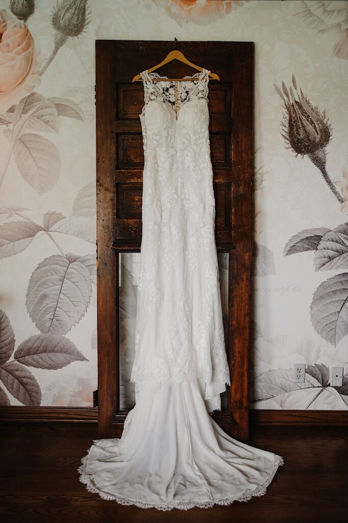 A lace wedding dress hanging on a wooden hanger on an antique door.  The door is leaning up against a wall with oversized flowers.