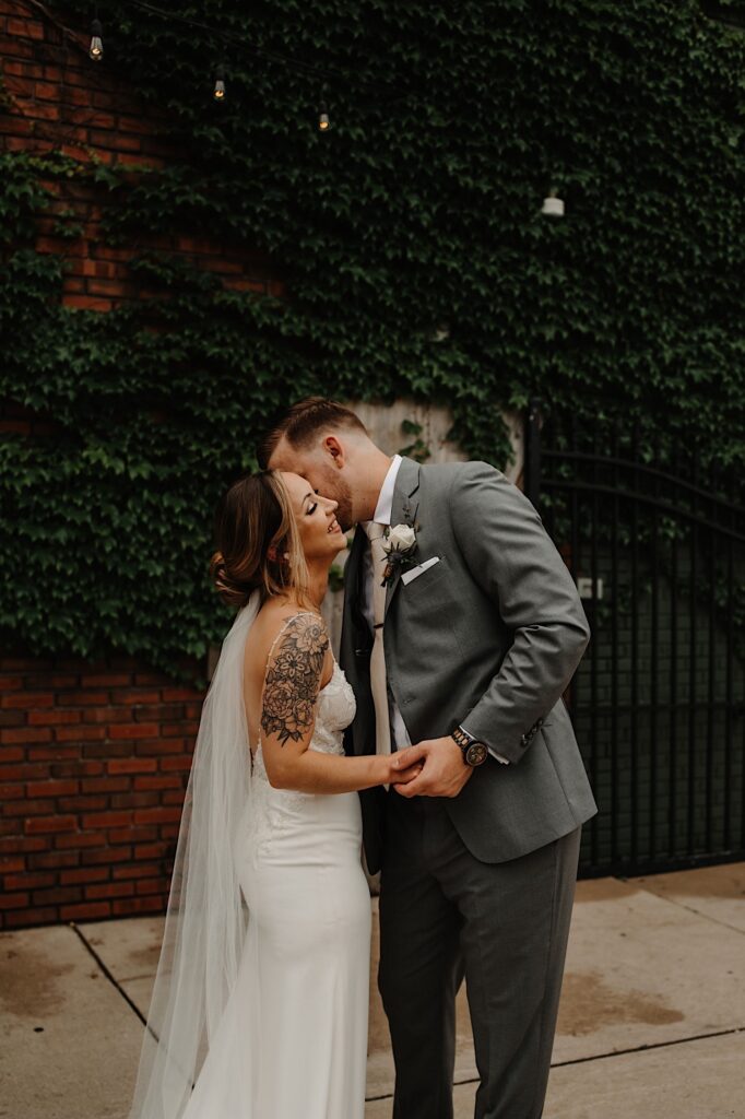 A groom kisses the bride on the cheek as she smiles outside of their wedding venue