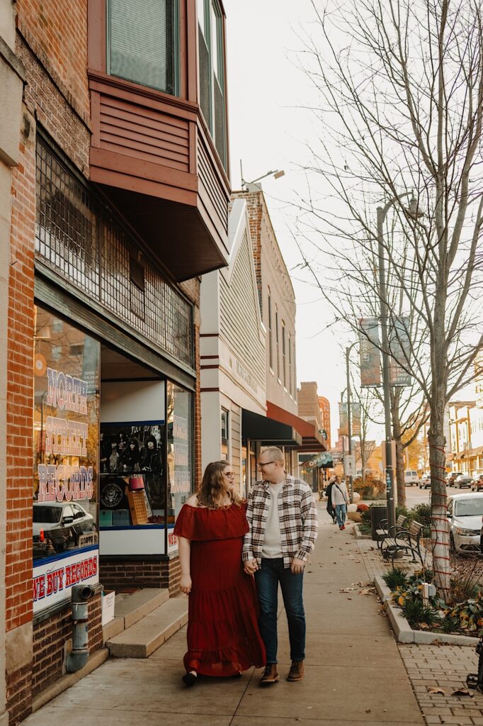 A couple holds hands walking down the street just outside of North Street Records smiling at one another during their Central Illinois engagement session.