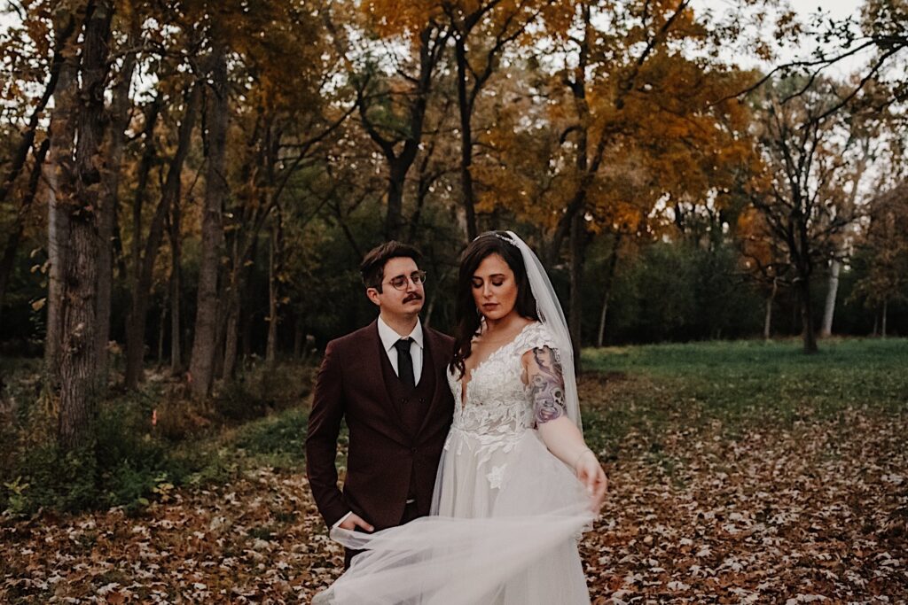 A groom in a maroon suit jacket and his bride in lace with tattoos on her arm pose for wedding portraits together surrounded by gorgeous fall colored leaves at their Chicagoland wedding venue.
