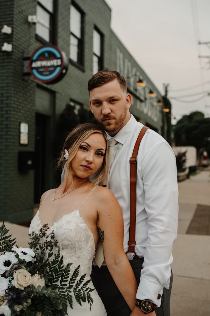 A bride and groom stand together and look at the camera while outside their wedding venue