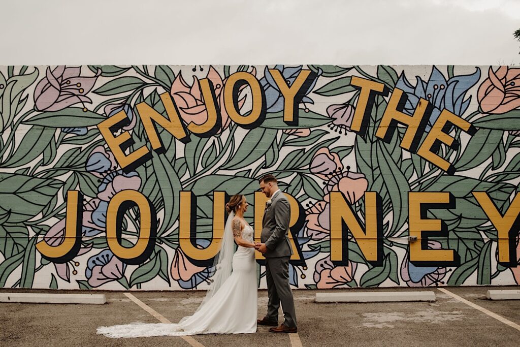 A bride and group stand facing one another and holding hands with a mural behind them reading "enjoy the journey" near their wedding venue the Ivy House