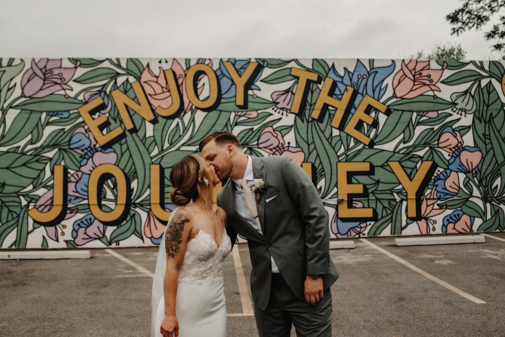 A bride and groom kiss one another and embrace with a mural behind them reading "enjoy the journey" near their wedding venue the Ivy House