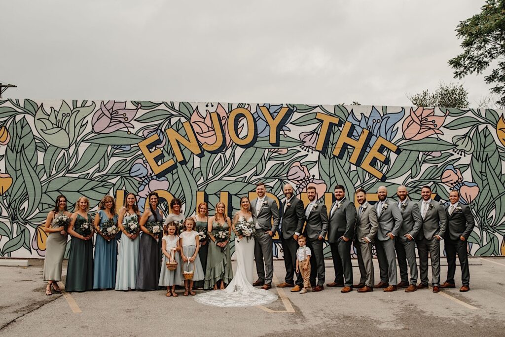 A bride and groom are joined by their wedding parties for a portrait with a mural behind them reading "enjoy the journey" near their wedding venue the Ivy House