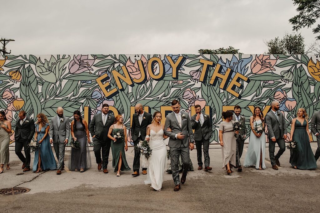 A bride and groom walk towards the camera together with their wedding parties doing the same on either side with a mural behind them reading "enjoy the journey" near their wedding venue the Ivy House