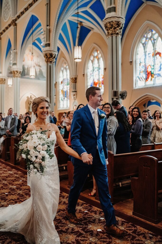 A bride and groom walk out of their ceremony holding hands and smiling at their guests at St Alphonsus Church.