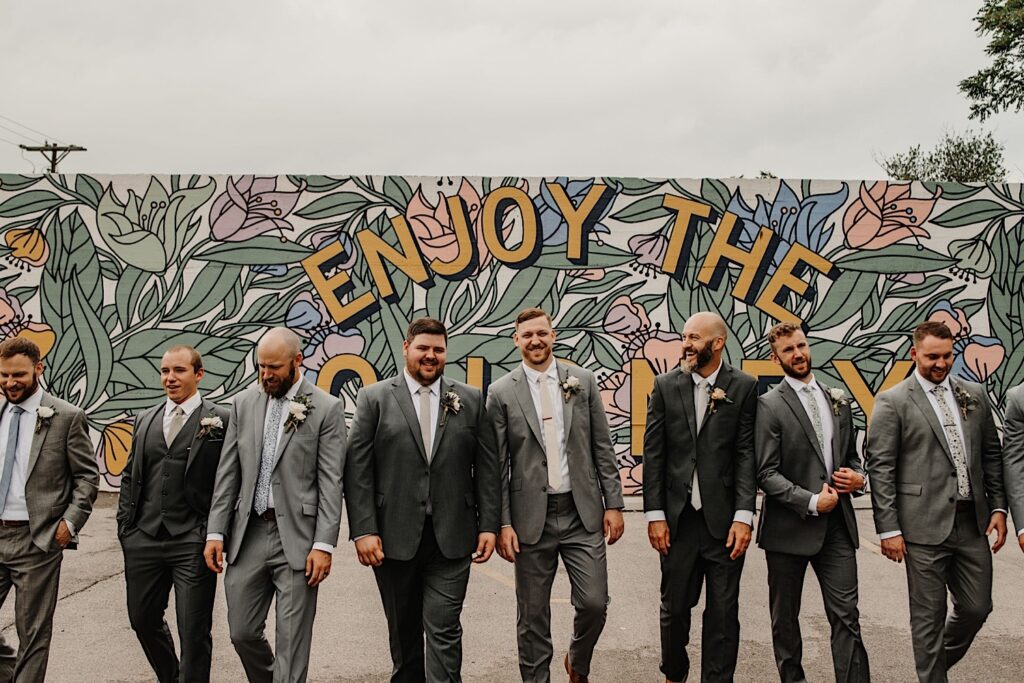 A groom walks towards the camera with his groomsmen as they laugh with a mural behind them reading "enjoy the journey" near their wedding venue the Ivy House