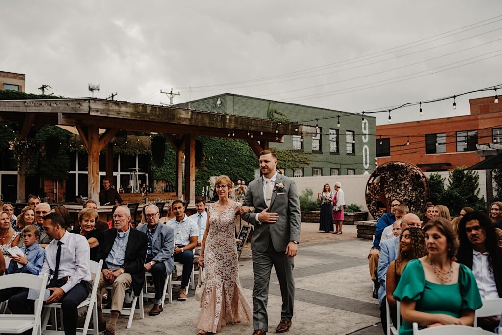 A groom is walked down the aisle by his mother with guests seated at the Ivy House's outdoor wedding space