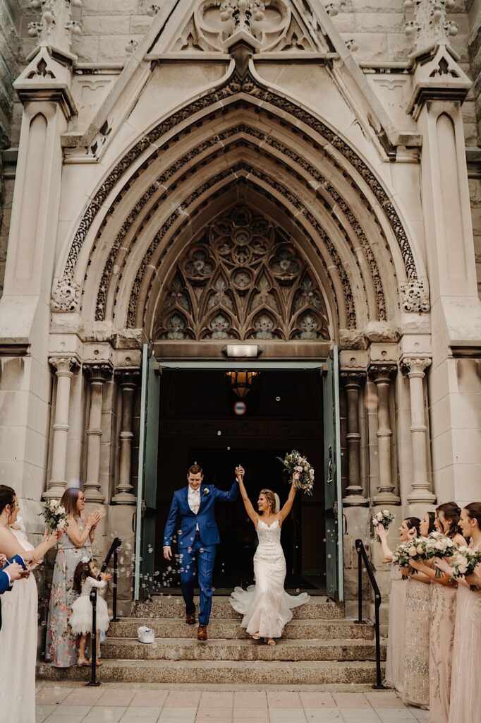The bride and groom walk out of St. Alphonsus Church in Chicago with their arms up in the air.  There are bubbles surrounding them and their guests are cheering.
