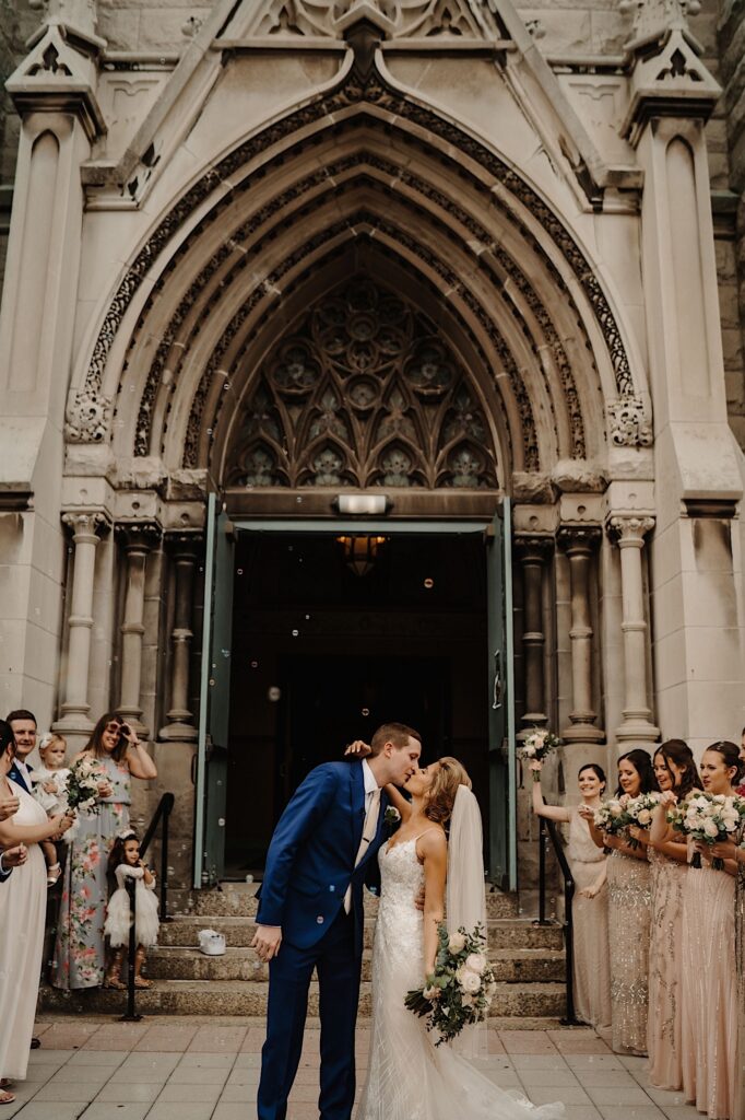 The bride and groom walk out of St. Alphonsus Church in Chicago with their arms up in the air.  There are bubbles surrounding them and their guests are cheering while they kiss.