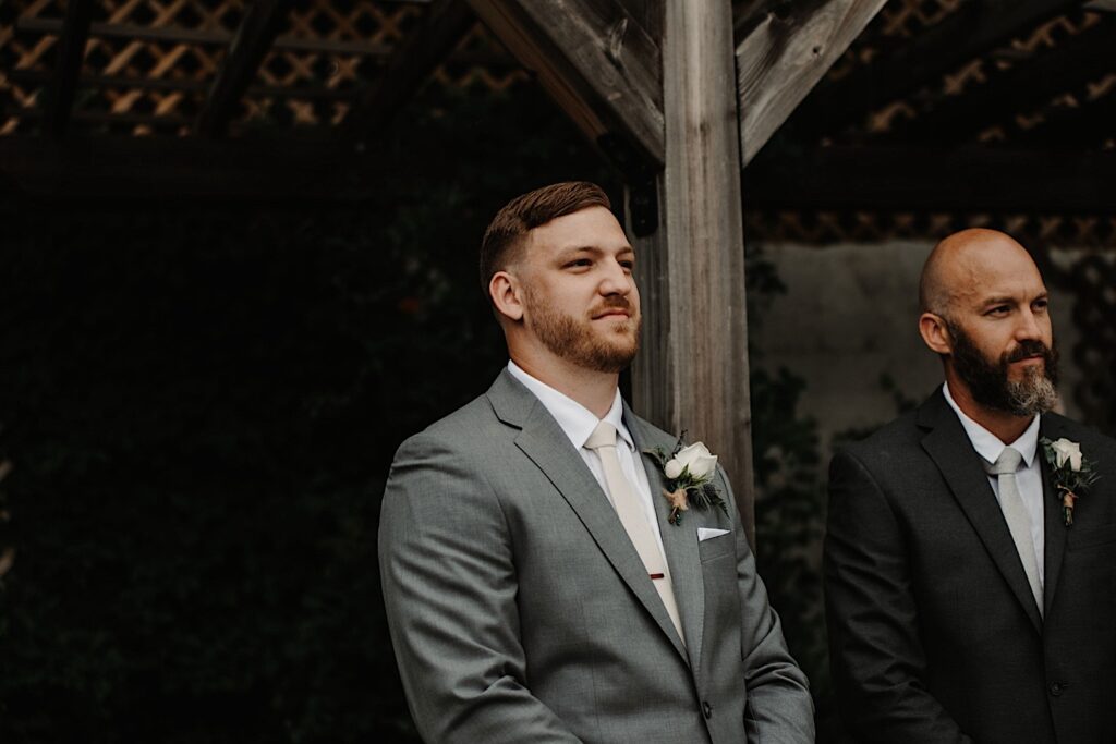A groom looks on with his best man at his side at his wedding ceremony in the Ivy House's outdoor space