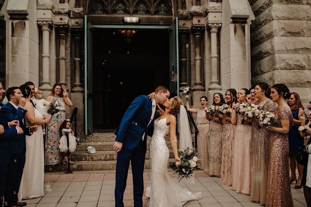 The bride and groom walk out of St. Alphonsus Church in Chicago with their arms up in the air.  There are bubbles surrounding them and their guests are cheering while they kiss.