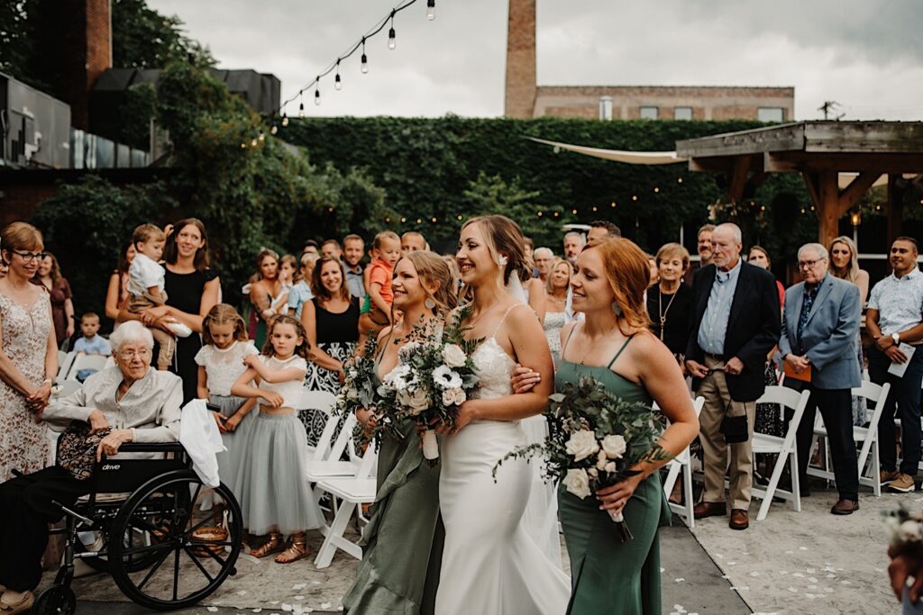 A bride is walked down the aisle of the Ivy House's outdoor ceremony space by her bridesmaids as guests stand and watch.