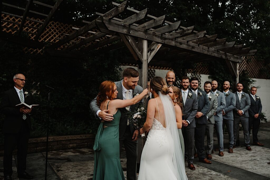 A groom is hugged by bridesmaids after they walked the bride down the aisle for the wedding ceremony at their outdoor wedding venue