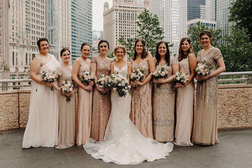 The bride stands with her bridesmaids on the Riverwalk in Chicago.  The bride wears a lace gown with a long train and the bridesmaids wear champagne colored dresses with beads.