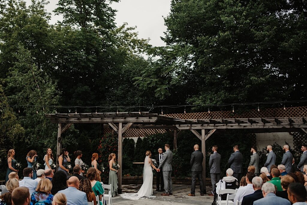 A bride and groom stand holding hands during their wedding ceremony at the Ivy House's outdoor ceremony space as their wedding parties and guests watch