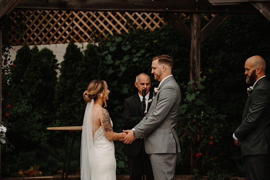 A bride and groom stand with one another and hold hands while their officiant speaks during their outdoor wedding ceremony.