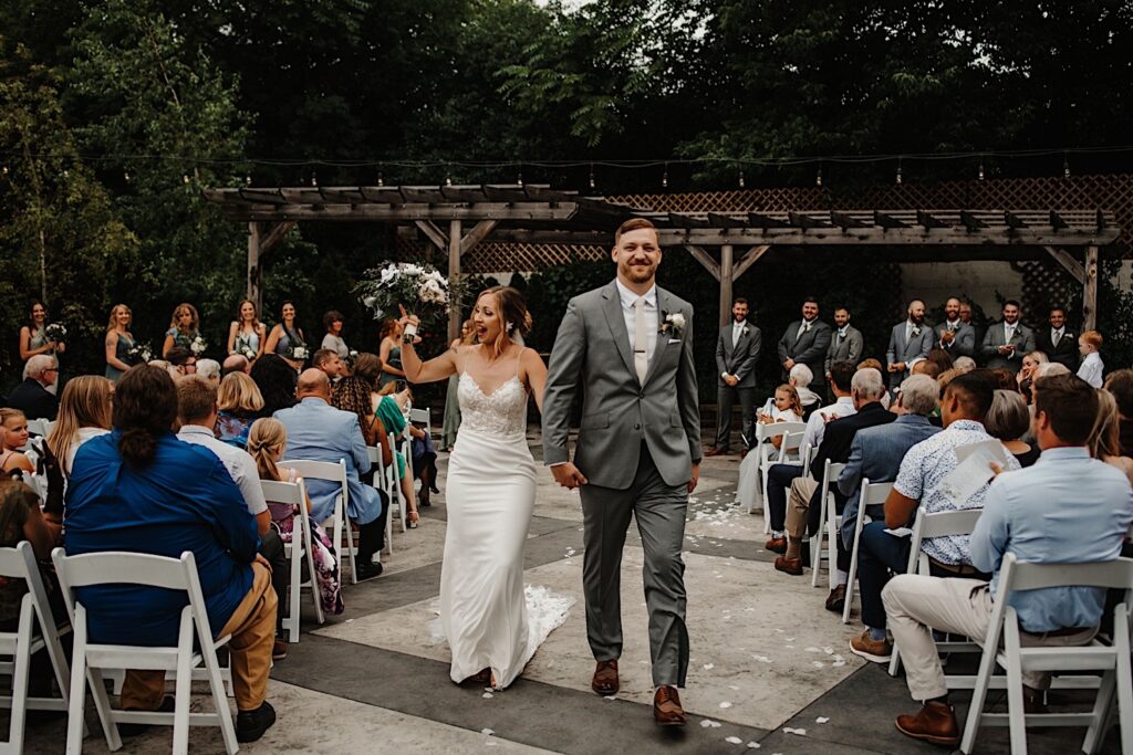A bride and groom walk down the aisle and celebrate together after their wedding ceremony at the Ivy House's outdoor ceremony space, guests cheer on either side of them