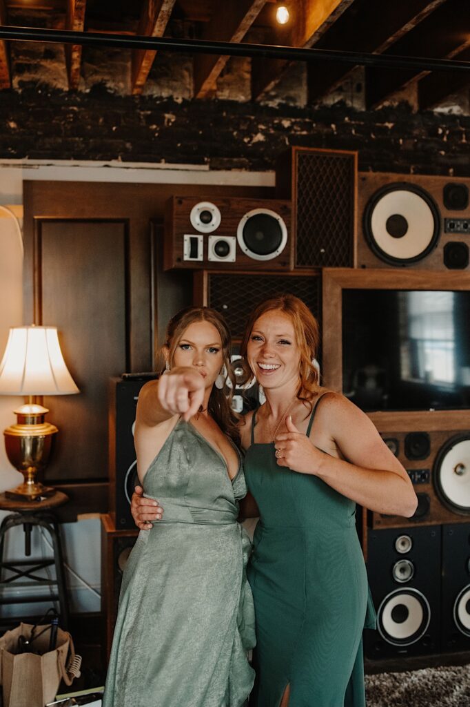 Two bridesmaids stand next to one another and pose for the camera, one points at the camera while the other gives a thumbs up