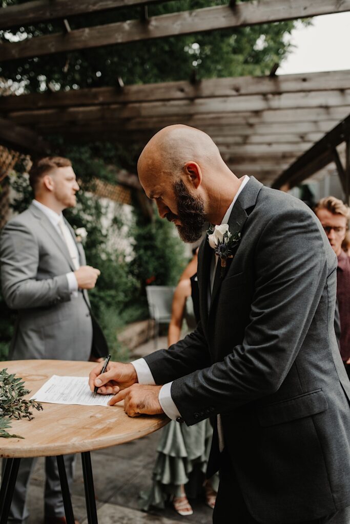 A groomsman signs as a witness on the wedding paperwork after his friend's outdoor wedding ceremony
