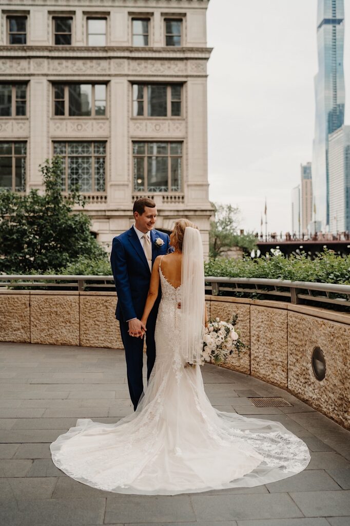 The bride stands with her groom with her the train of her wedding dress spread out around her.  They smile at each other as they take wedding portraits in Chicago.