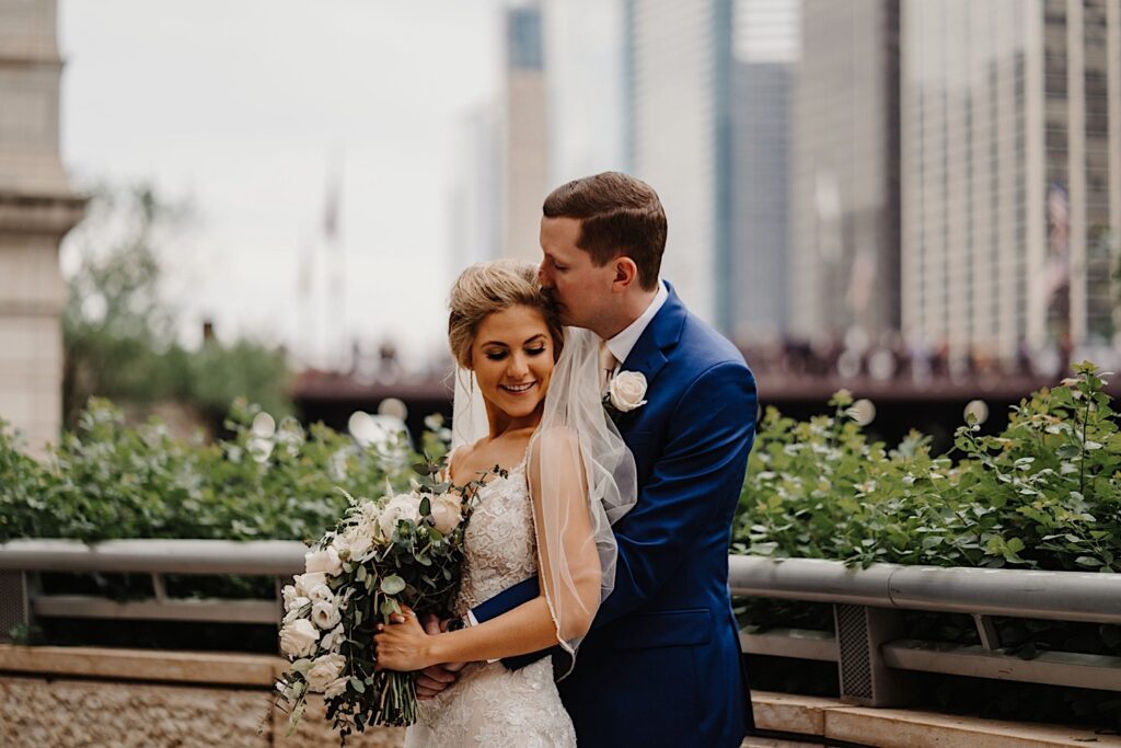 The bride stands with her back to the groom as he wraps his arms around her waste and kisses her temple.  She smiles while looking at the ground, you can see a Chicago bridge in the background of their wedding portraits.