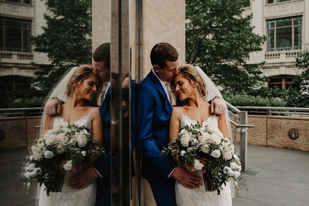 A bride and groom stand next to a glass door holding one another close, you can see their reflection in the other half of the image.