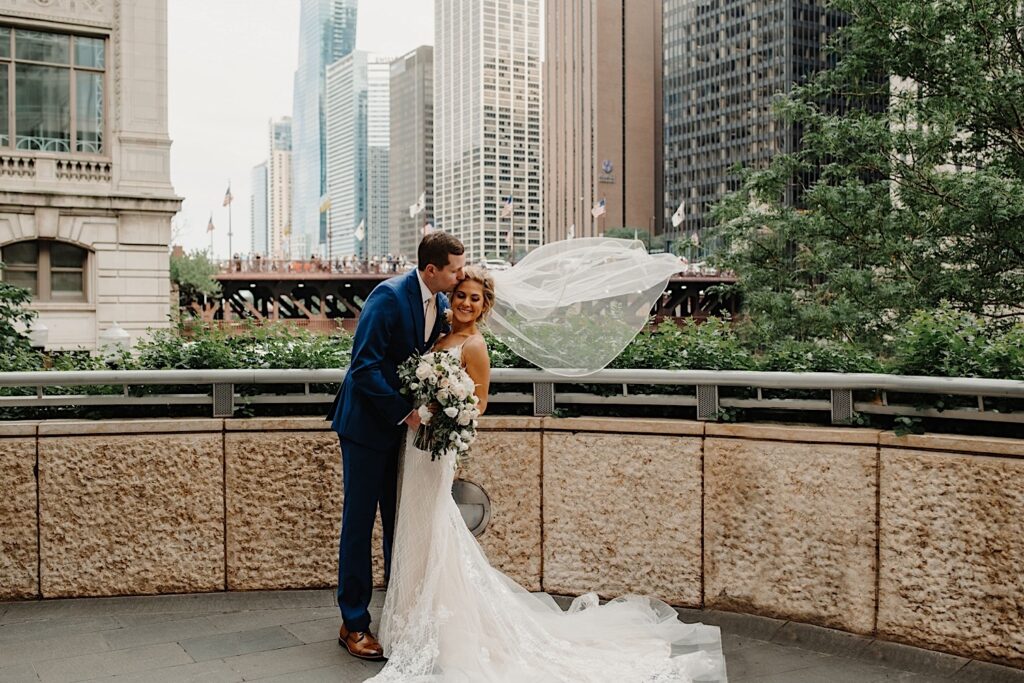 The groom kisses the brides' temple on a bridge of the Chicago Riverwalk.  The brides veil floats in the breeze.