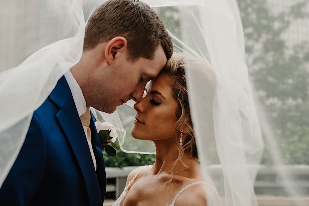 The bride and groom share an intimate moment together with their foreheads pressed together and their eyes closed.  They're tucked underneath the brides veil
