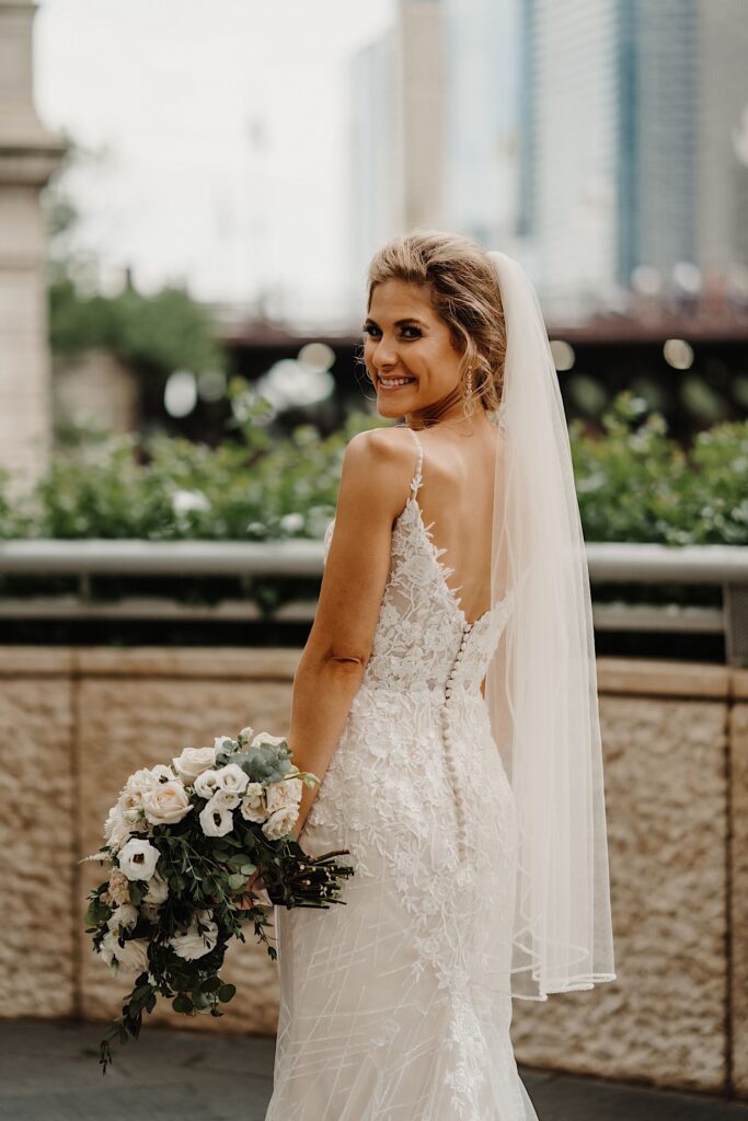 The bride smiles while looking over her shoulder during her wedding portraits on the Chicago Riverwalk.  She wears a lace dress with spaghetti straps and a white veil.