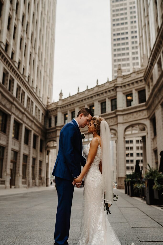 The bride and groom stand in front of the Tribune Tower in Chicago with their foreheads pressed together.  They hold hands and take a moment together.