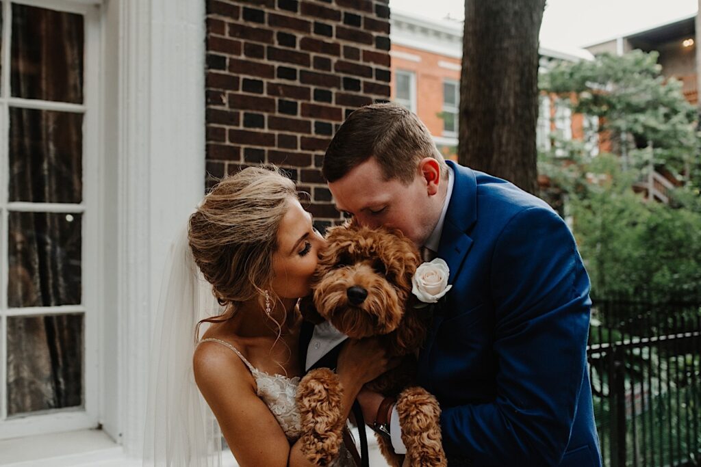 A bride and groom hold their dog between them as they both kiss his head during their wedding portraits