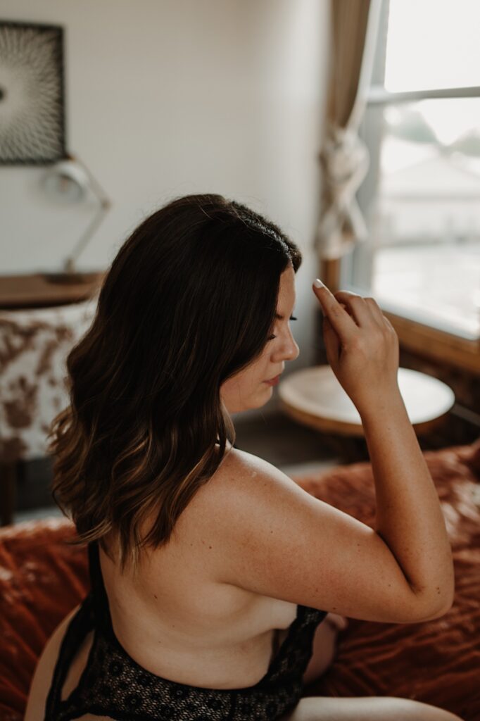 A woman sits profile to the camera on a bed with a pink velvet bedspread in a Wicker Park apartment in Chicago.  She is wearing a black lace one piece for her Chicago boudoir session.  This image is closer to the subject.
