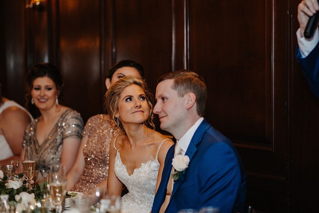 A bride sits next to the groom and looks up as a guest gives a speech during their wedding reception at Salvatore's in Chicago