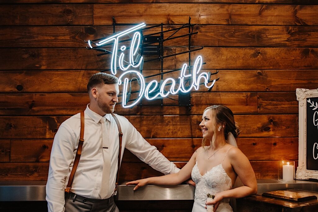 A bride and groom stand next to one another and smile at a bar during their wedding reception at Ivy House, between them is a neon sign that reads "Til Death"