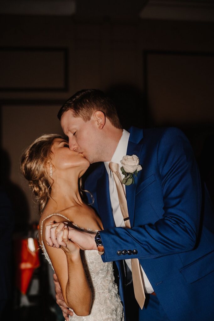 A bride and groom kiss in a dimly lit room as they hold hands