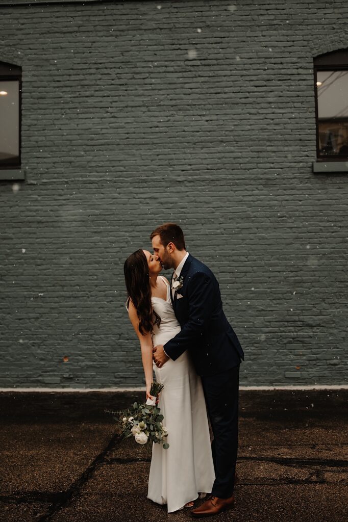 A bride and groom kiss while standing in the snow in front of a green painted  brick wall.