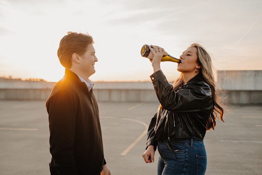 A fiancée drinks champagne out of the bottle while looking at her fiancé during their engagement session.  They are standing on a parking garage in Bloomington Normal.