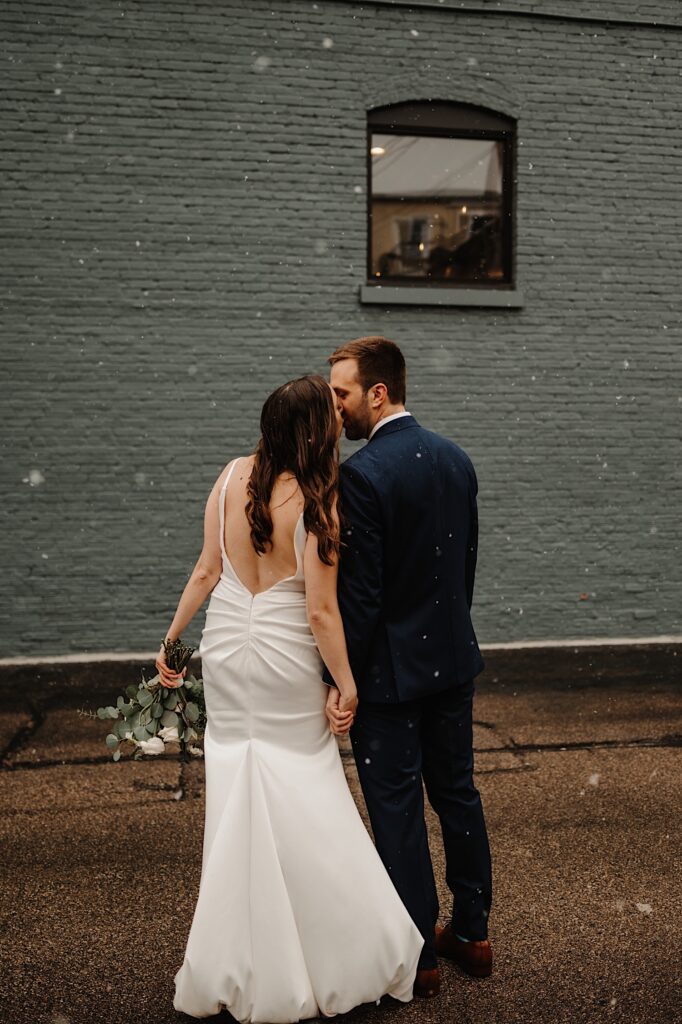 A bride and groom kiss while standing in the snow in front of a green painted  brick wall facing away from the camera