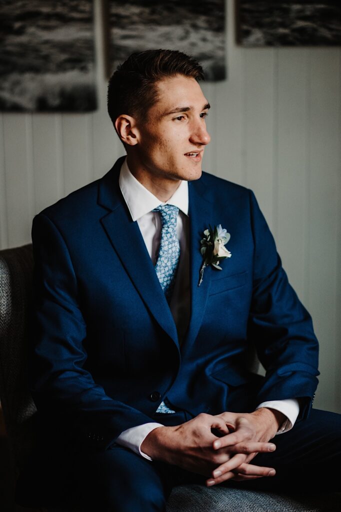 The groom sits in the getting ready space of his Wisconsin wedding venue.  He is wearing a blue suit with a blue floral tie and a white and blue boutonnière.  