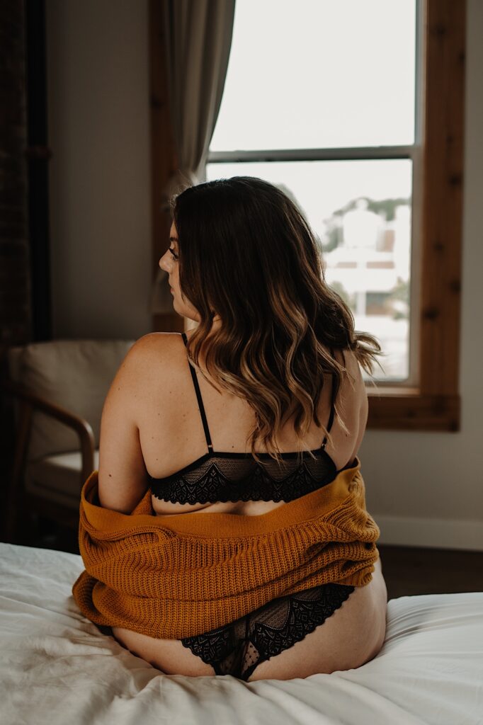 A woman sits on the edge of her bed in a matching two piece lace lingerie set with a chunky orange sweater wrapped around her.