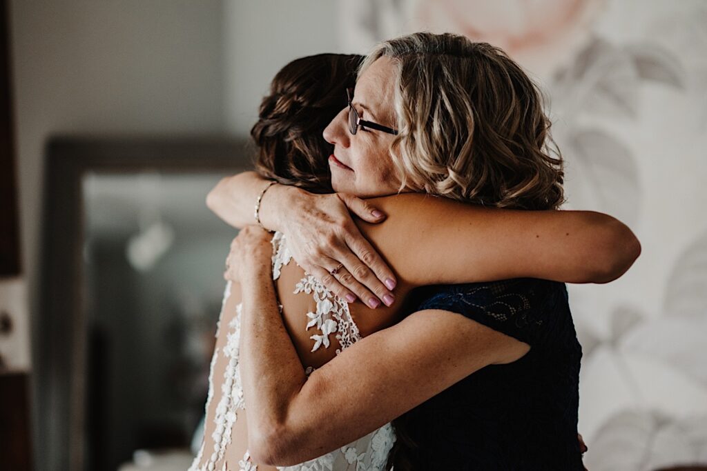 A bride hugs her mom in the getting ready room before her wedding day.  She wears a lace dress and her mom wears a dark blue dress.