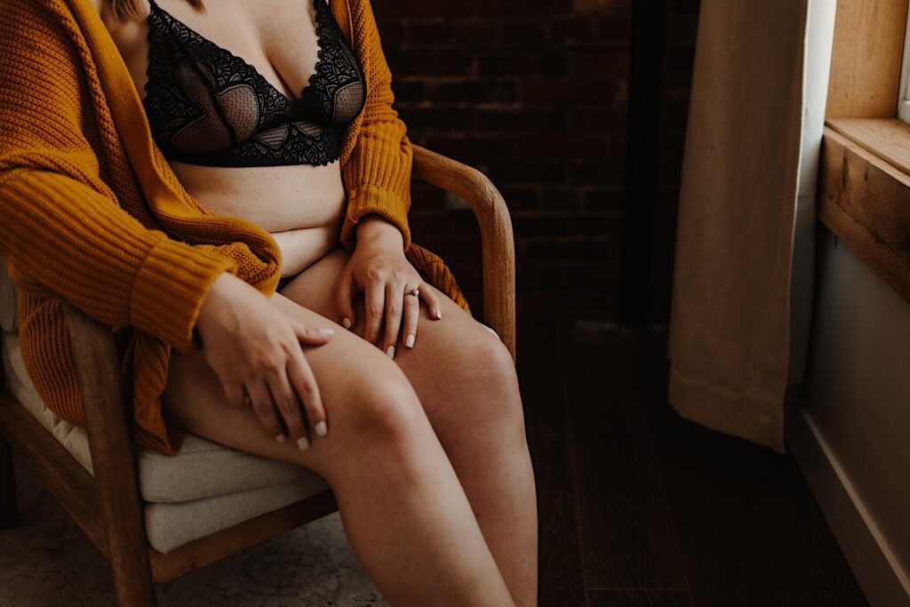 A woman sits on a chair looking out the window with her hands sitting on her lap.  She is wearing a matching lace set of lingerie and a chunky knit orange sweater.